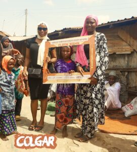 A group of women and children stand together during a charity clothing drive in Offa, Kwara State, holding a frame labeled "Cloth Bank" as part of Colgar's outreach initiative.